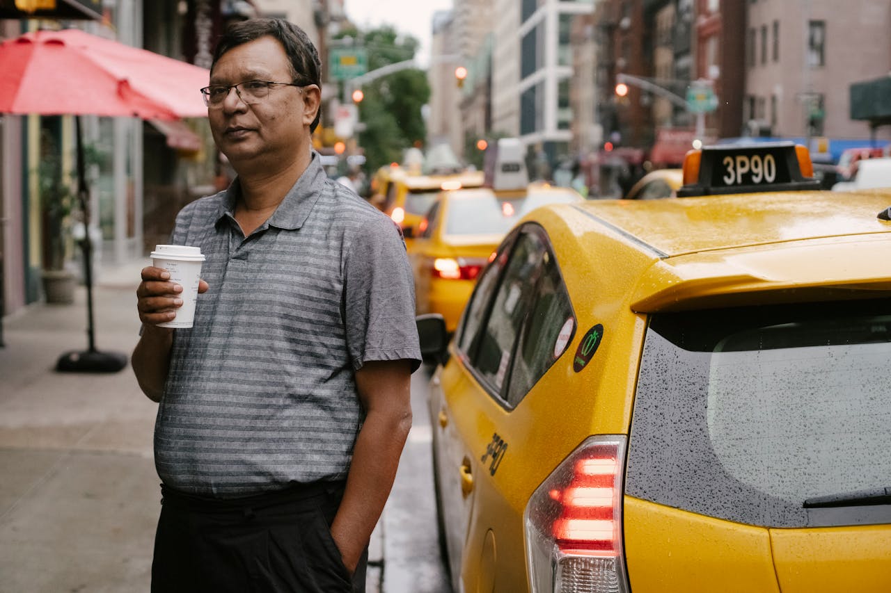 Calm Asian middle aged male in eyeglasses standing on roadside with cup of hot drink to go near yellow cab with hand in pocket